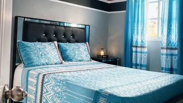 A bedroom with a bed, a nightstand, a lamp, a window with curtains, and an air conditioning unit.