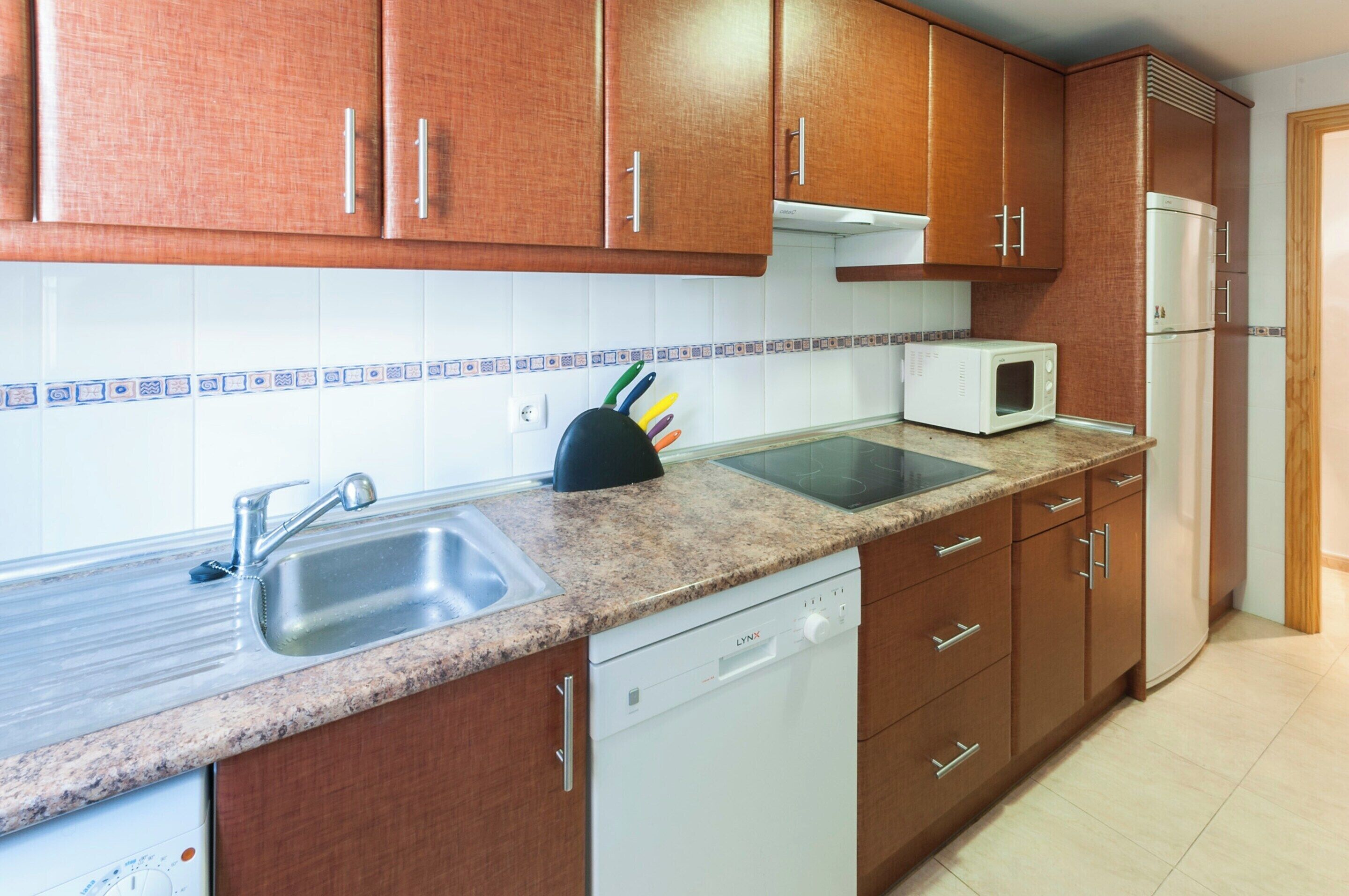 Una cocina con armarios de madera, encimera de granito, pileta, lavavajillas, microondas y refrigerador.