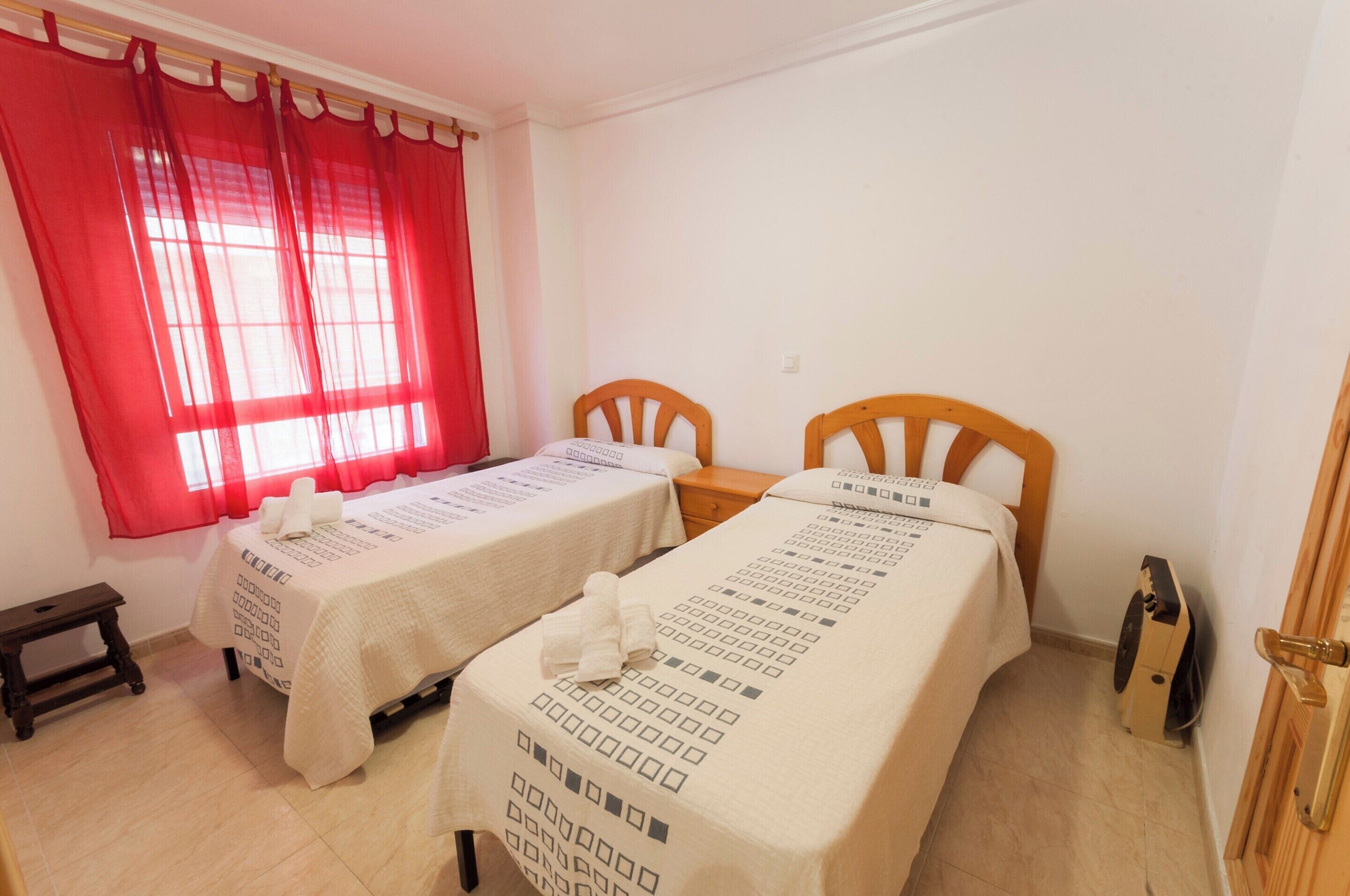 A hotel room with two beds, a small table, a chair, a window with red curtains, and a door.