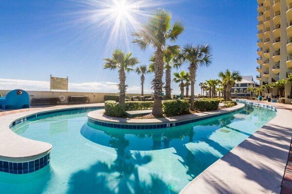 A swimming pool with a curved design, surrounded by palm trees and a tall building in the background.