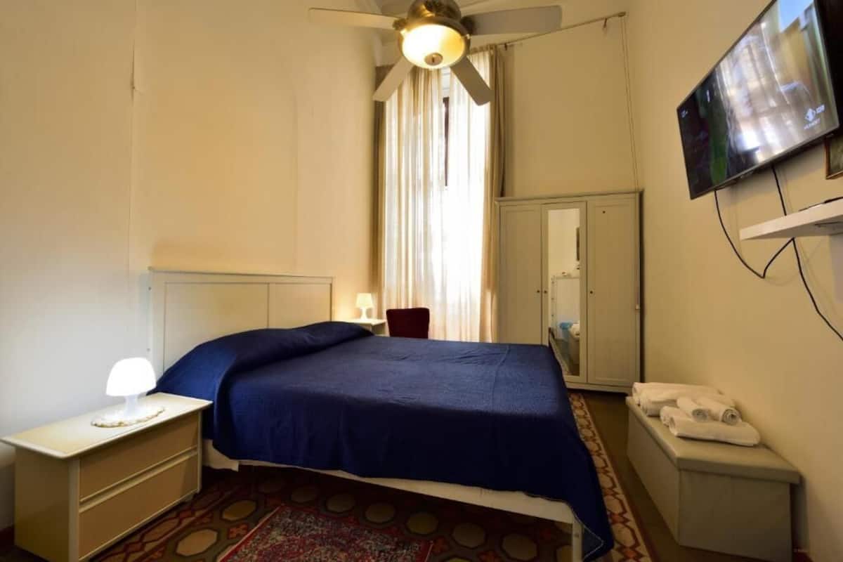 Habitación de hotel con cama, mesita de noche con lámpara, televisor montado en la pared, armario y un pequeño banco con toallas.