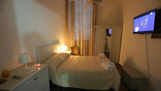 Habitación de hotel con cama, lámpara de noche, televisor y una pequeña mesa con lámpara y control remoto.