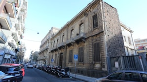 Una calle con coches y motocicletas estacionados, un edificio de fachada de piedra y un cielo azul despejado.