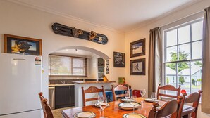 A dining area with a wooden table set for four, a kitchen in the background, and a window with curtains. - Willow Lodge - Ohakune Holiday Home (Ohakune)