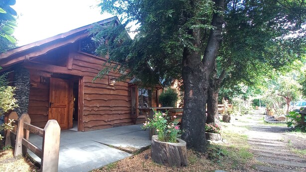A rustic log cabin with a wooden porch, surrounded by lush greenery and trees. - Cabañas Rucapillan (Panguipulli)