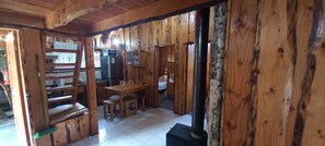 A wooden cabin interior with a dining table, chairs, a kitchen area with appliances, and a wood-burning stove.