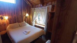 A wooden cabin bedroom with a bed, two wall-mounted lamps, a window with curtains, and a wooden shelf. - Cabañas Rucapillan (Panguipulli)