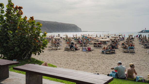 Ein Strand mit vielen Liegestühlen und Sonnenschirmen, eine Rasenfläche mit Bänken und eine Klippe im Hintergrund.