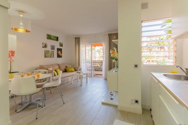 Un salon moderne avec un canapé, une table basse et un coin repas. La cuisine est équipée d’un évier et d’une fenêtre donnant sur des immeubles.