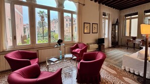 A spacious room with large windows, red upholstered chairs, a small table with a vase, and a white sofa. - Monty Don's secret garden on the Grand Canal (Venezia)