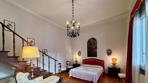 A bedroom with a wooden staircase, a chandelier, a bed with a red headboard, a red and white patterned rug, and a wooden chair. - Monty Don's secret garden on the Grand Canal (Venezia)