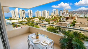 A balcony with a table set for two, overlooking a cityscape with high-rise buildings and mountains. - Apartamentos Velas al Mar - by SOHO (Santa Marta)