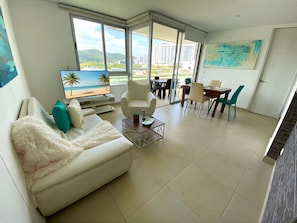 A modern living room with a white sofa, a coffee table, and a TV displaying a beach scene. There is a dining area with a table and chairs, and large windows offering a view of buildings and hills. - Apartamentos Velas al Mar - by SOHO (Santa Marta)