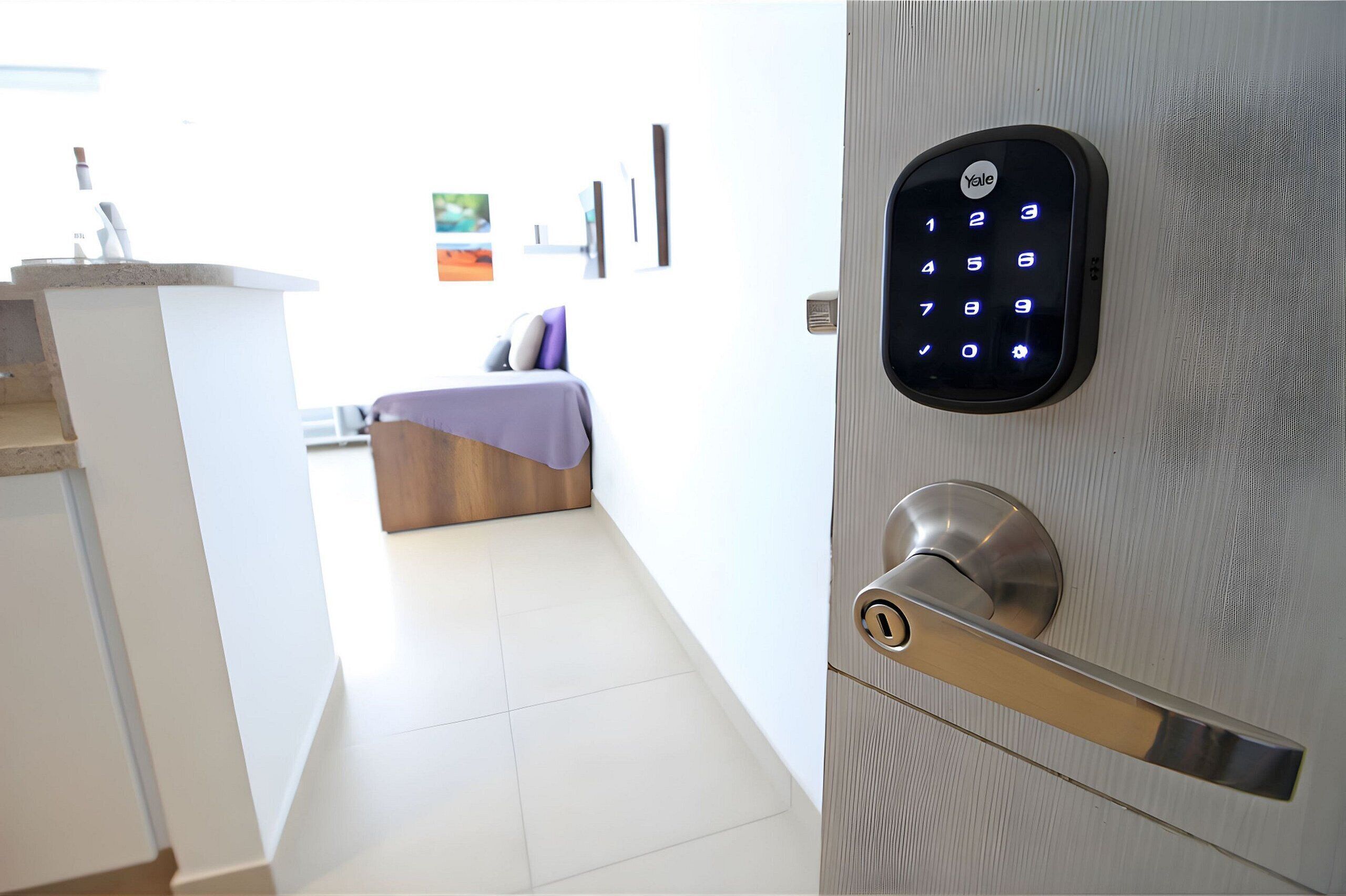 A modern hotel room with a Yale smart lock on the door, a bed with a purple blanket, and a small desk with a vase.