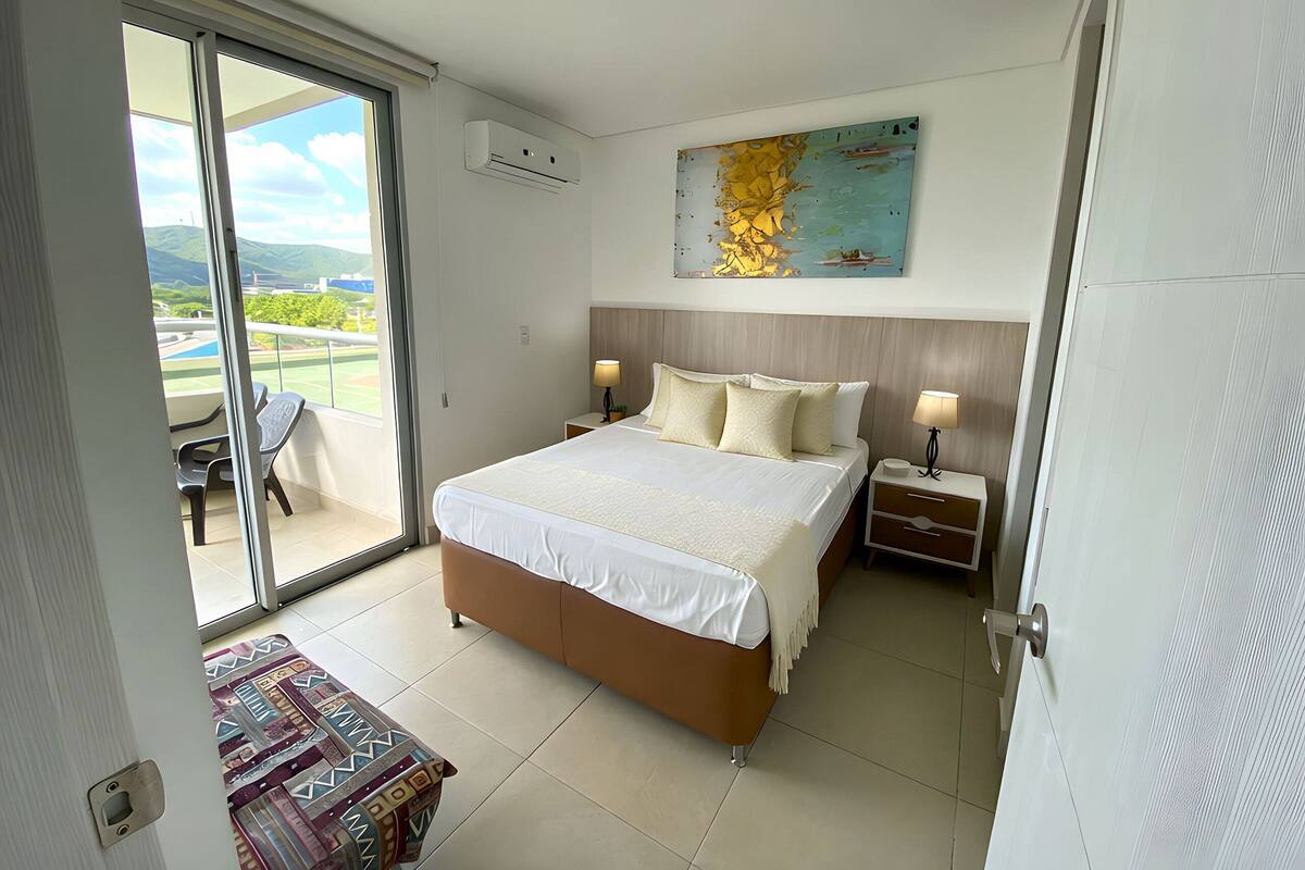 A hotel room with a bed, bedside tables, a window with a view of mountains, and a painting on the wall.
