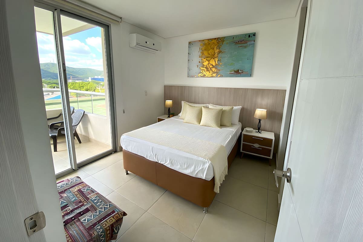 A hotel room with a bed, bedside tables, a window with a view of mountains, and a painting on the wall.
