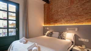 Ein ordentlich bezogenes Bett mit aufgerollten Handtüchern, eine Ziegelwand, ein Fenster mit Vorhängen und ein Nachttisch mit Buch und Fernbedienung.