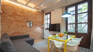 Ein modernes Wohnzimmer mit Backsteinwand, einer Couch, einem Glastisch mit Stühlen, einem Fernseher und einem großen Fenster mit Blick auf Bäume.