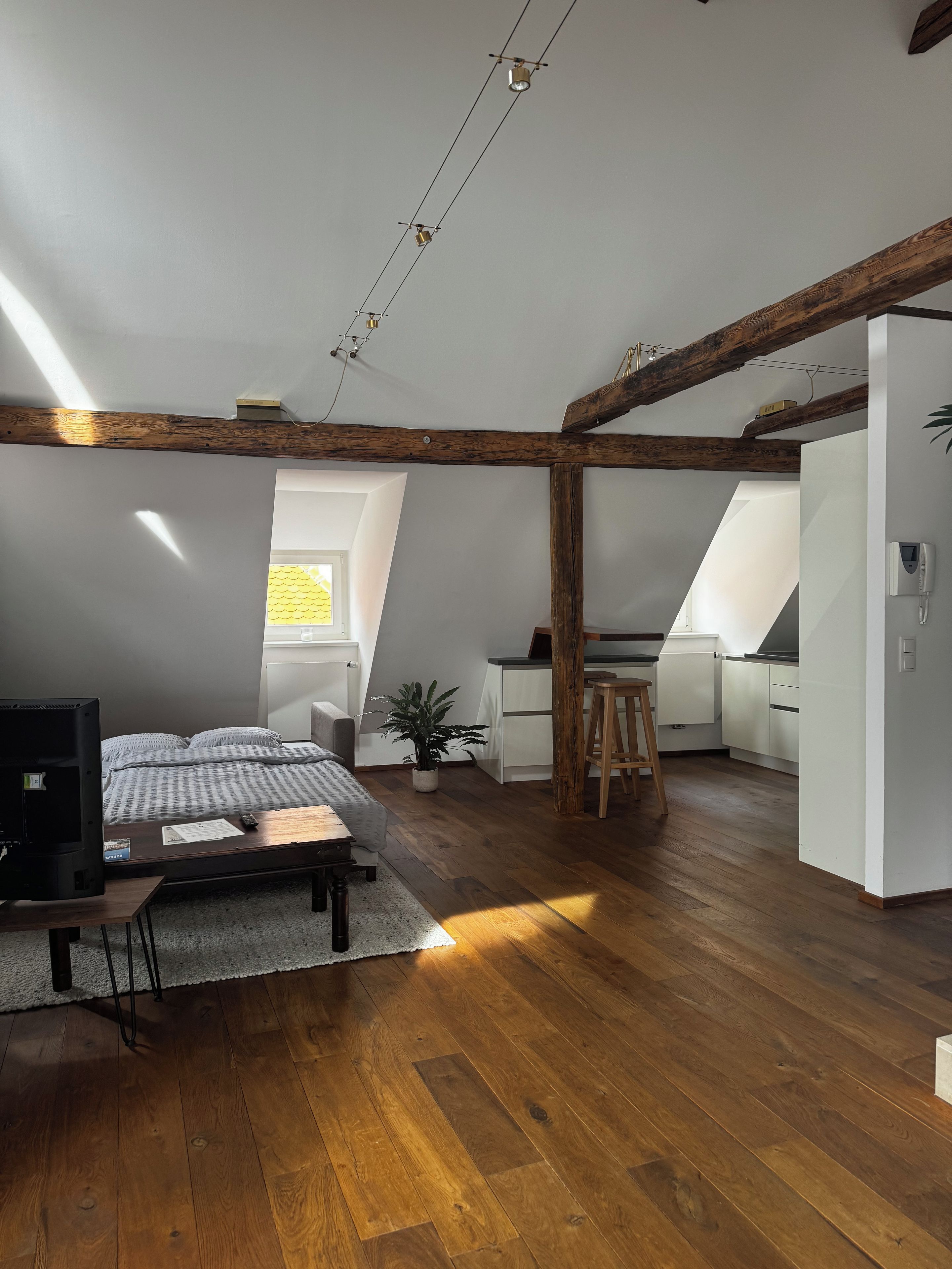 A loft-style room with a wooden floor, a bed, a desk, and a kitchenette.