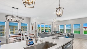 A modern kitchen with a large island, white countertops, and a stainless steel sink. There are multiple windows with blinds, a ceiling fan, and a view of a green landscape. - Beachfront Condo • Rooftop Pool • Sleeps 8 (Mexico Beach)
