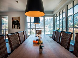 Un comedor con una mesa de madera larga, sillas de mimbre, una planta en maceta y un plato de frutas.
