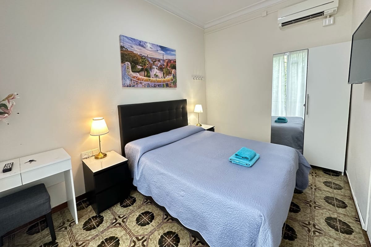 A hotel room with a bed, bedside tables, a desk, a wardrobe, and an air conditioning unit.