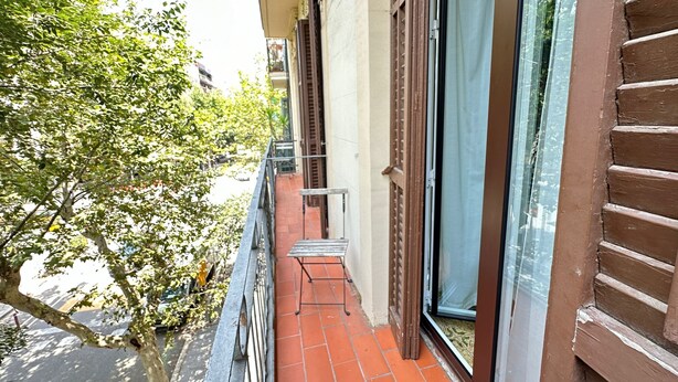 Ein schmaler Balkon mit Metallgeländer, roten Ziegelböden und großen Fenstern mit Holzläden.
