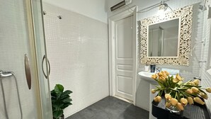 Ein Badezimmer mit Duschabtür aus Glas, einer weißen Fliesenwand, einem Waschbecken mit dekorativem Spiegel und einer Topfpflanze.