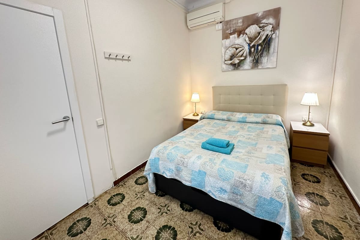 A hotel room with a bed, two bedside tables with lamps, an air conditioning unit, a door, and a patterned floor.