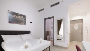 Una camera d'albergo con un letto, un quadro, una porta e un bagno visibile attraverso una porta aperta.
