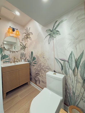 Une salle de bain avec un meuble-lavabo en bois, des toilettes blanches et une fresque murale représentant des plantes tropicales.