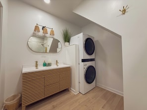 A modern bathroom with a wooden vanity, a round mirror, a wall-mounted light fixture, and a built-in washer and dryer.