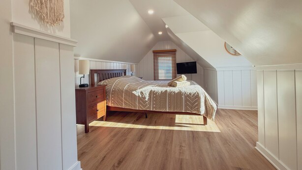 Ein Schlafzimmer mit geneigter Decke, einem Bett mit gemustertem Überwurf, einem hölzernen Nachttisch, einem an der Wand montierten Fernseher und einem Wandbehang aus Makramee.
