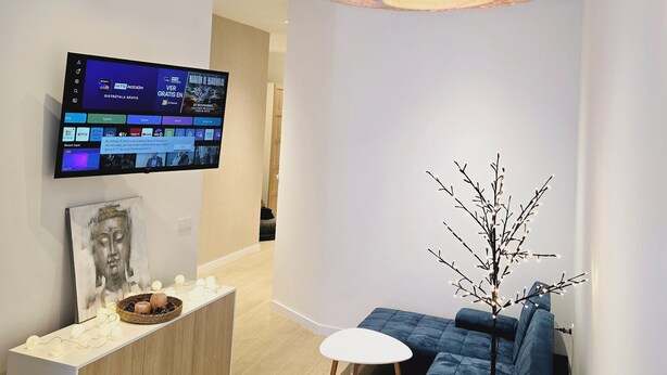 Ett modernt vardagsrum med en platt-TV som är monterad på väggen, en blå soffa, ett litet vitt bord och en dekorativ gren med ljus.