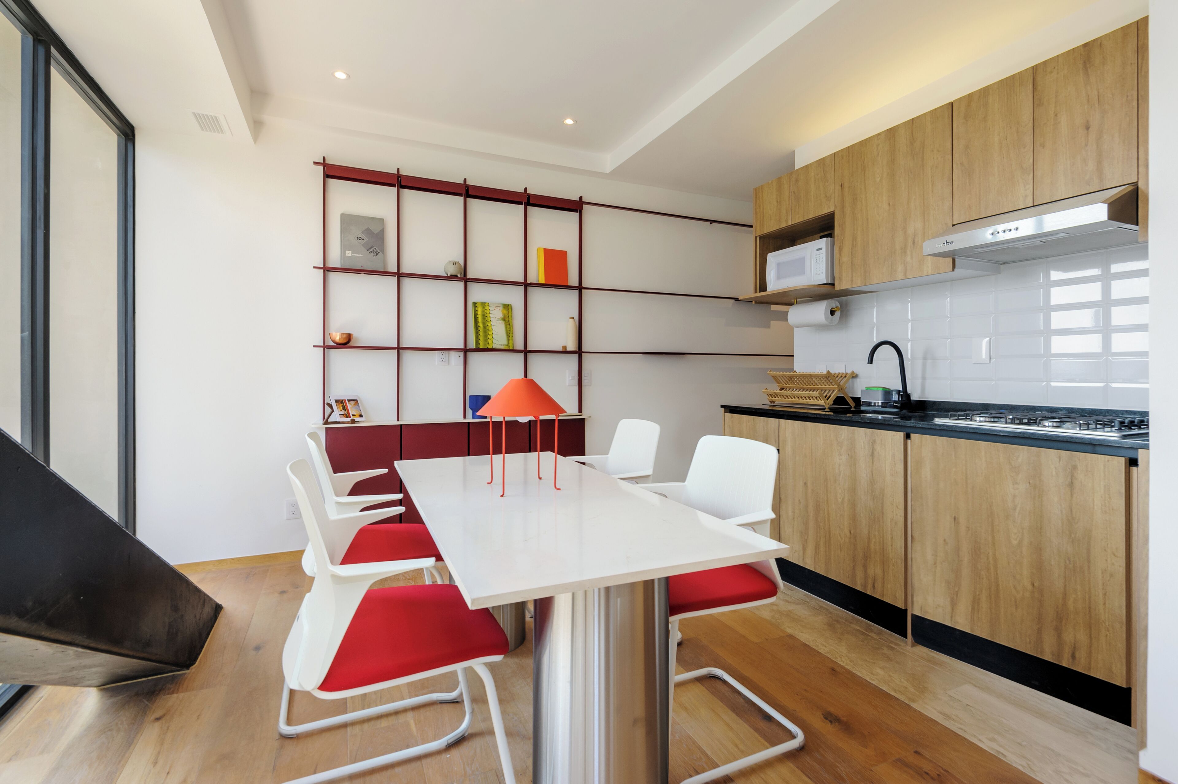 Una cocina moderna con armarios de madera, encimera blanca y estufa integrada. Hay un comedor con mesa blanca y sillas rojas.