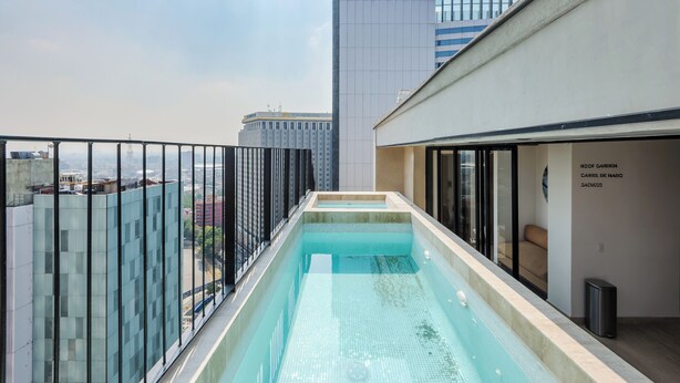 Una piscina en la azotea con barandilla de cristal, un rascacielos al fondo y un cartel que indica "ROOF GARDEN, CARLISLE DE NADO, JACUZ 25".