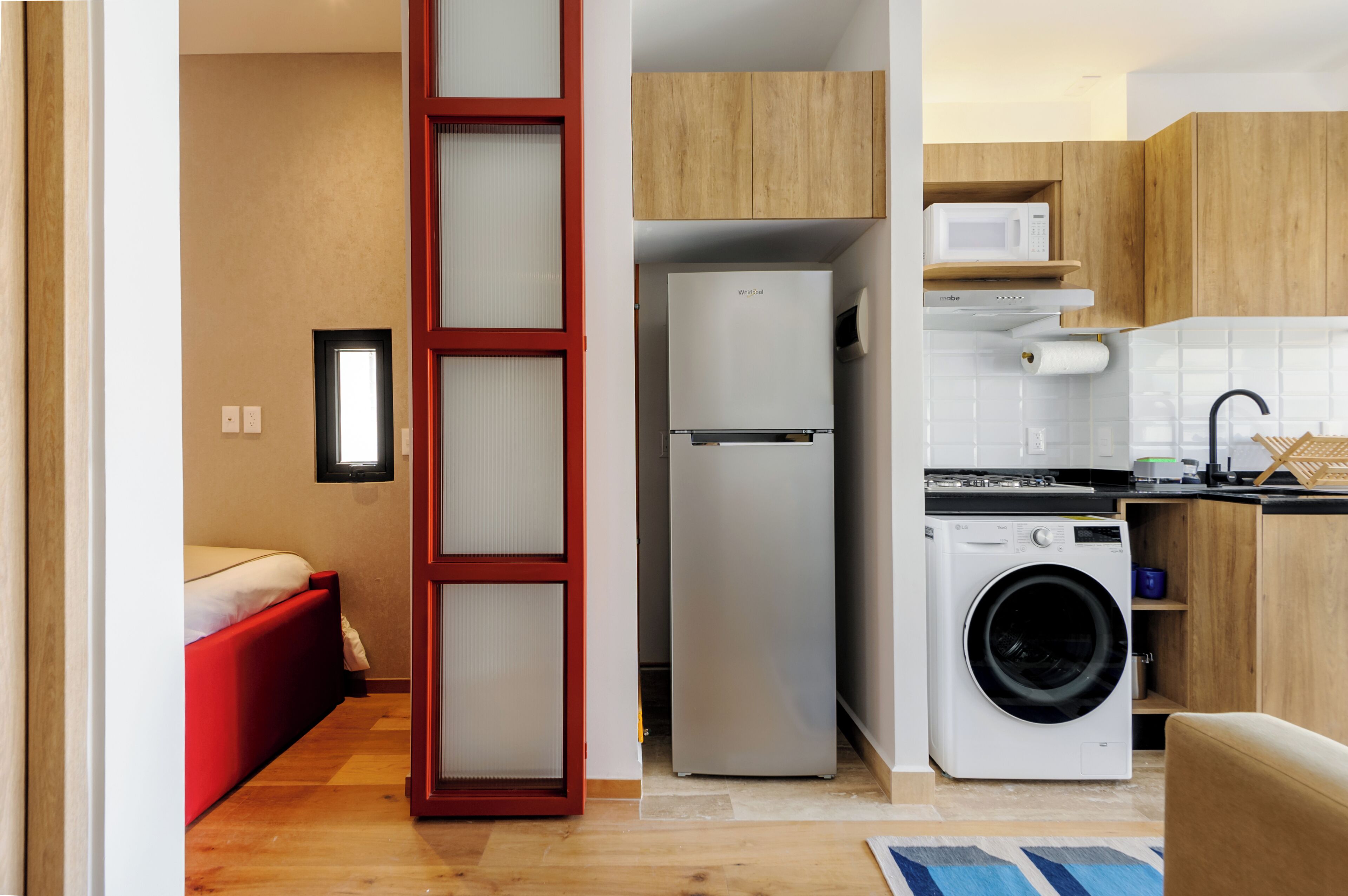 Un espace de vie compact comprenant un coin cuisine, un lave-linge et un lit avec une tête de lit rouge.