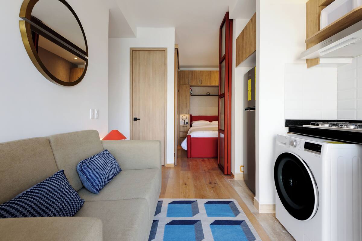 A compact living space with a sofa, a washing machine, a bed, and a small kitchen area.
