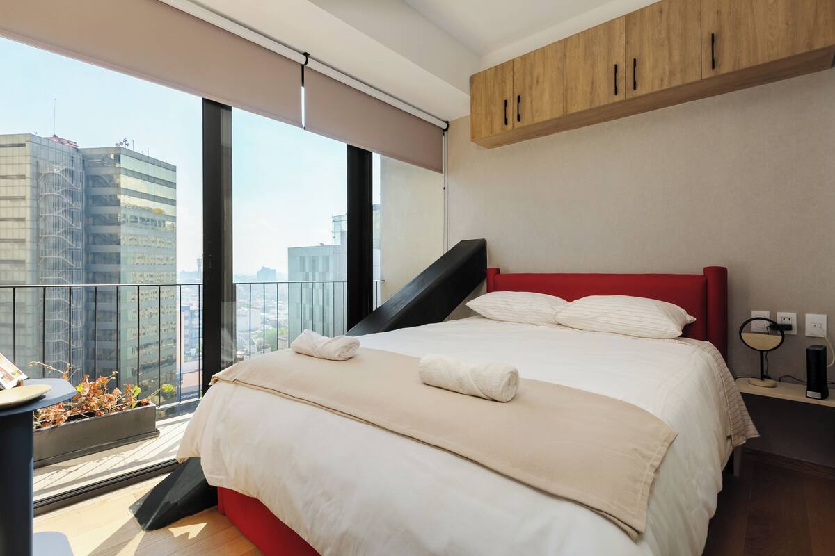 A modern hotel room with a large bed, a red headboard, a small bedside table with a lamp, and a view of the city through a large window.