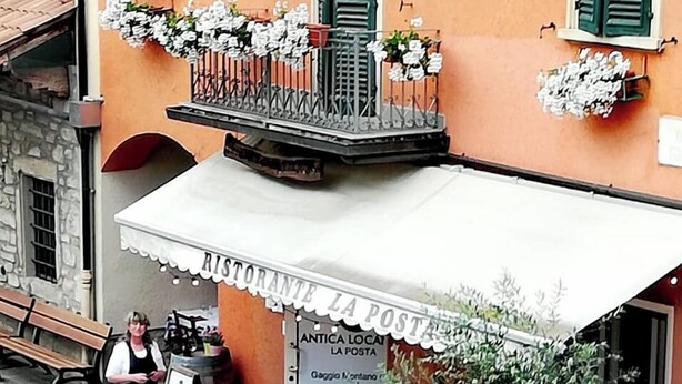 Un área de estar al aire libre con mesas y sillas, un letrero que dice "Ristorante La Posta" y un balcón con flores.