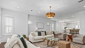 A modern living room with a white sofa, armchairs, a glass coffee table, and a wooden side table. There is a chandelier hanging from the ceiling.