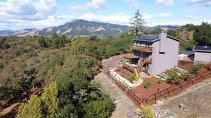 A modern house with a balcony, surrounded by a fenced garden and a mountainous backdrop. - Charming 3 BD Retreat - Spa, EV Charger, Views (Santa Rosa)