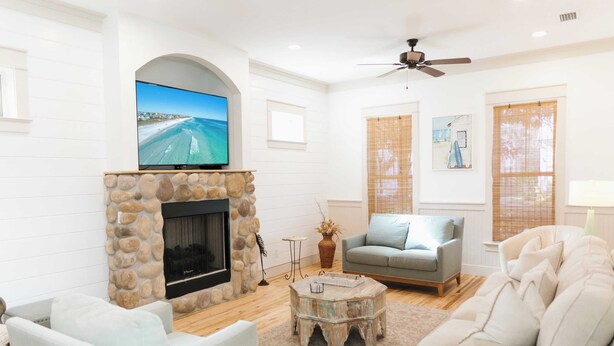 A living room with a stone fireplace, a flat-screen TV mounted above it, and a ceiling fan. - 5 Mins to Beach, Shared Pool, Covered Deck, Bikes Heron Cottage by Avantstay (Santa Rosa Beach)