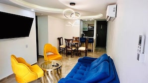 Uma sala de estar moderna com um sofá azul, cadeiras amarelas, uma mesa de centro de vidro e uma televisão de tela plana fixada na parede.