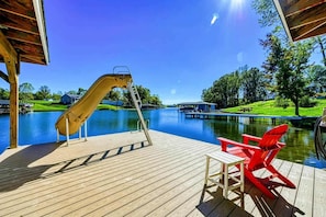 A wooden deck with a slide, red chairs, and a table overlooking a lake.