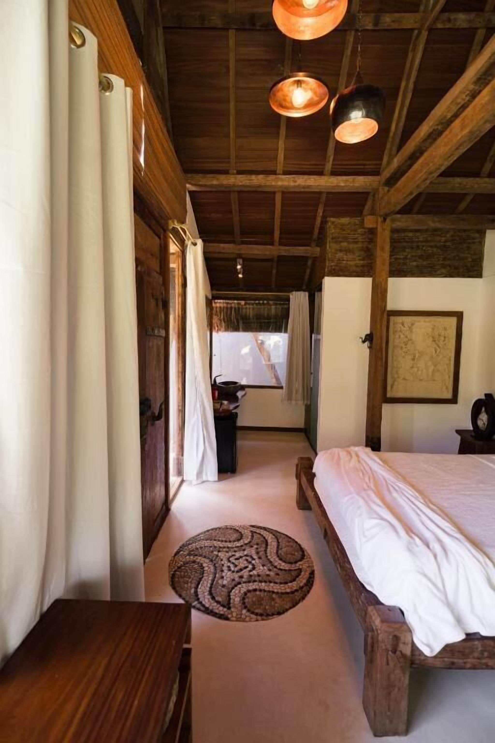 A rustic bedroom with a wooden bed, a round woven rug, and a view of the outdoors through a window.