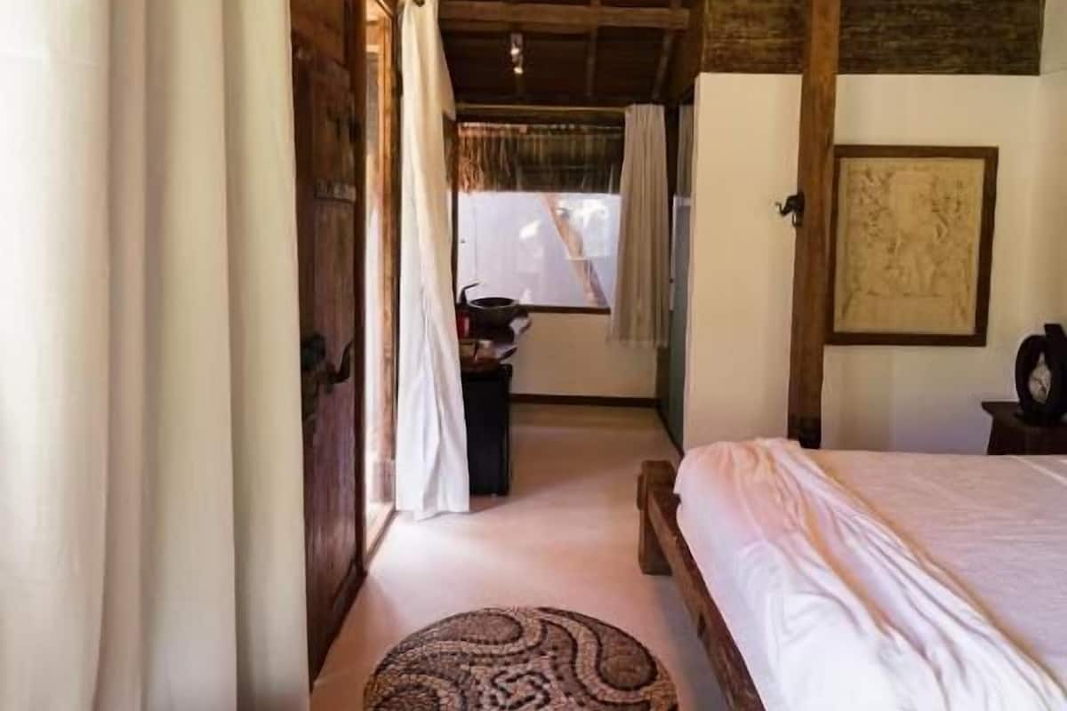 A rustic bedroom with a wooden bed, a round woven rug, and a view of the outdoors through a window.