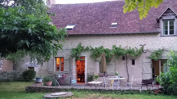 A rustic stone building with a tiled roof, a garden with potted plants, and outdoor furniture.