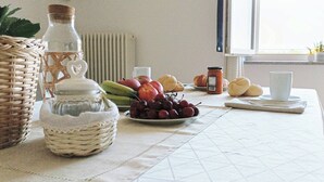 A dining table with a white tablecloth, a wicker basket, a glass jar, a plate of fruits, and a window with a view of greenery. - Quick Home Mimosa - One Bedroom Apartment, Sleeps 4 (Alice Castello)