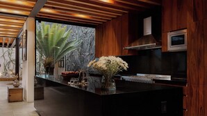A modern kitchen with a large island, wooden ceiling beams, a stone backsplash, and a view of a tropical plant. - Casa Carmen Xólotl (Oaxaca)